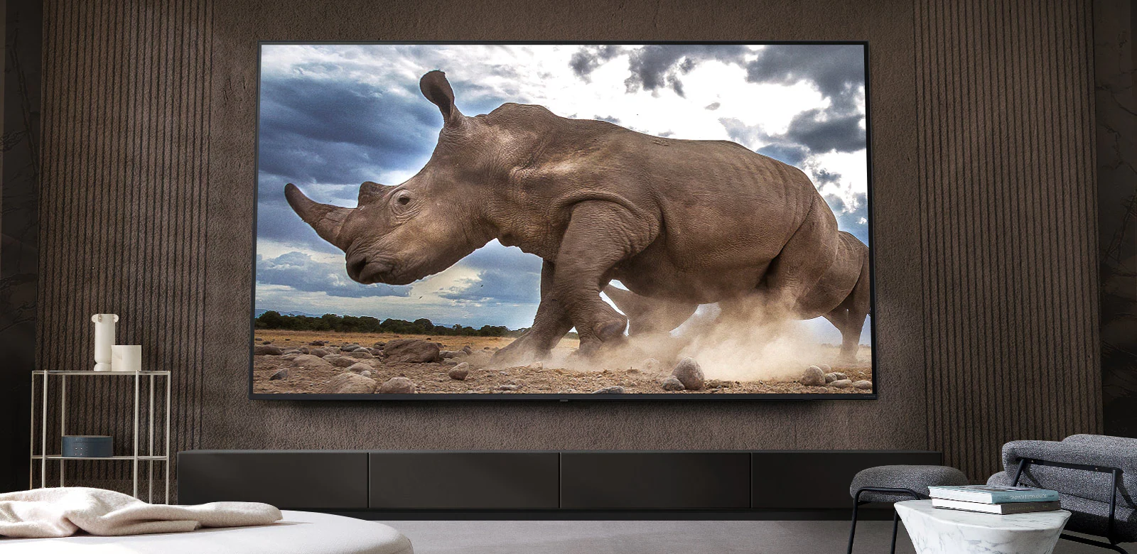 Носорог в обстановке сафари демонстрируется на сверхбольшом телевизоре LG TV, установленном на коричневой стене гостиной, окруженной модульной мебелью кремового цвета.