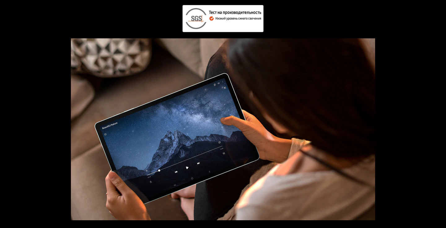 Женщина на диване держит обеими руками Galaxy Tab S9&nbsp;FE+ и смотрит полноэкранное видео в горизонтальном режиме. Логотип SGS с надписью &laquo;Показатели проверены. Низкое излучение синего света&raquo;.