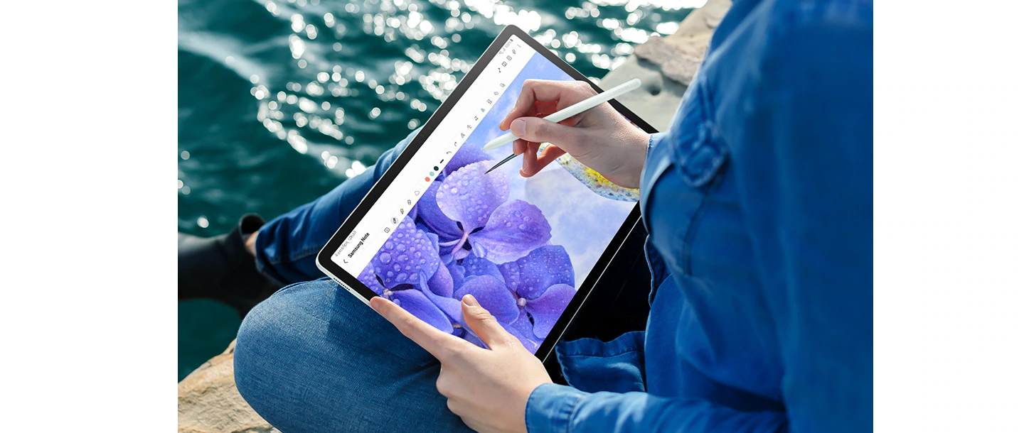 Человек, сидящий у воды, держит в руках Galaxy Tab S9&nbsp;FE+ в горизонтальном режиме и пером S&nbsp;Pen рисует цветок в открытом на экране приложении Samsung Notes.
